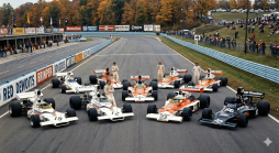 Formel-1-Rennwagen und Fahrer der 70er Jahre auf einer Rennstrecke, umgeben von Herbstbäumen und Zuschauern.