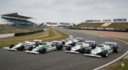 Fünf klassische Williams Formel-1-Rennwagen, weiß und grün, auf einer Rennstrecke mit Zuschauertribüne und Werbetafeln unter bewölktem Himmel.