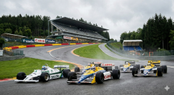 Historische F1-Rennwagen, darunter Williams und Brabham, fahren auf einer nassen Strecke in Spa-Francorchamps mit Tribünen und Wald im Hintergrund.