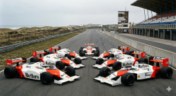 Acht McLaren Formel-1-Rennwagen auf der Rennstrecke Zandvoort, umgeben von Dünen und Tribünen, präsentiert in einer Formation.