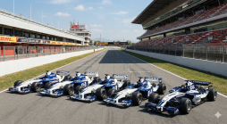 Sieben historische Williams F1 Rennwagen, blau-weiß lackiert, auf der Start-Ziel-Geraden des Circuit de Catalunya unter blauem Himmel.
